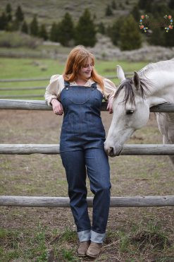 a woman wearing feminine denim overalls stands at a fence with a horse behind her