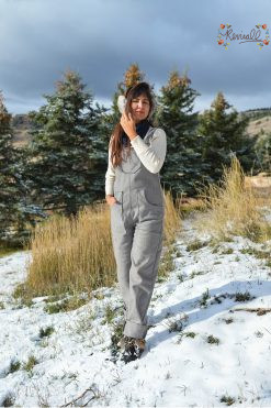 a woman wearing a pair of grey wool American Made overalls walks on a hillside of snow with mountains in the background