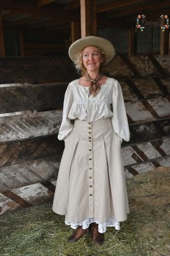 a western woman stands in a horse barn wearing a wool skirt and cowgirl hat