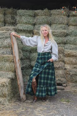 a woman stands in front of a hay pile. She is wearing a blue and green plaid cotton skirt with ruffle and a striped prairie blouse with big sleeves.