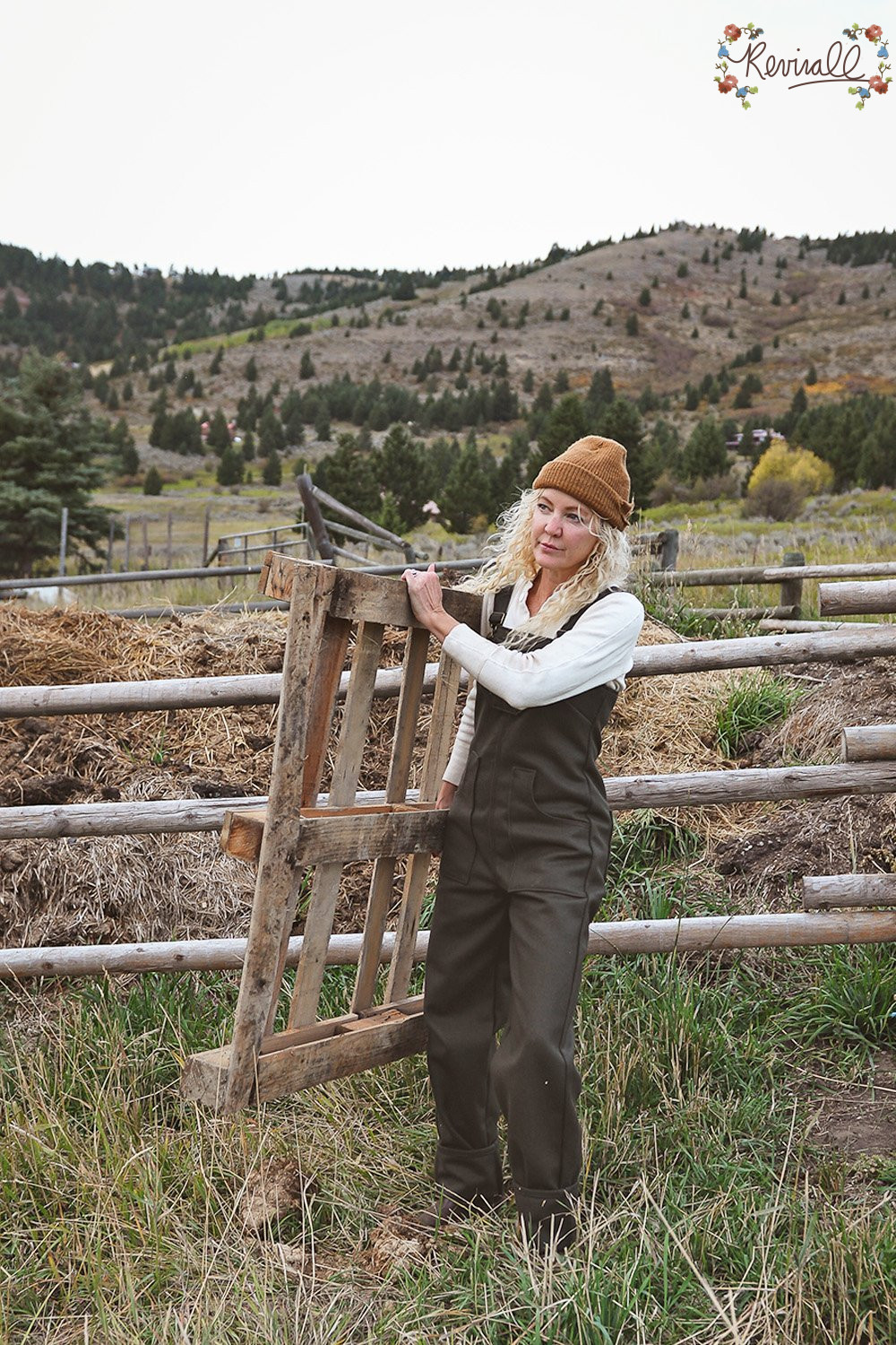 a woman wearing green wool overalls carries a pallet with a wooden fence line and mountains behind her.