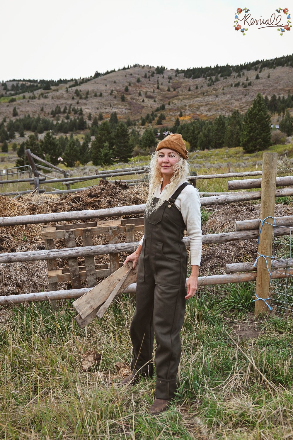 a woman wearing green wool overalls carries a pallet with a wooden fence line and mountains behind her.