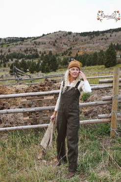 a woman wearing green wool overalls carries a pallet with a wooden fence line and mountains behind her.