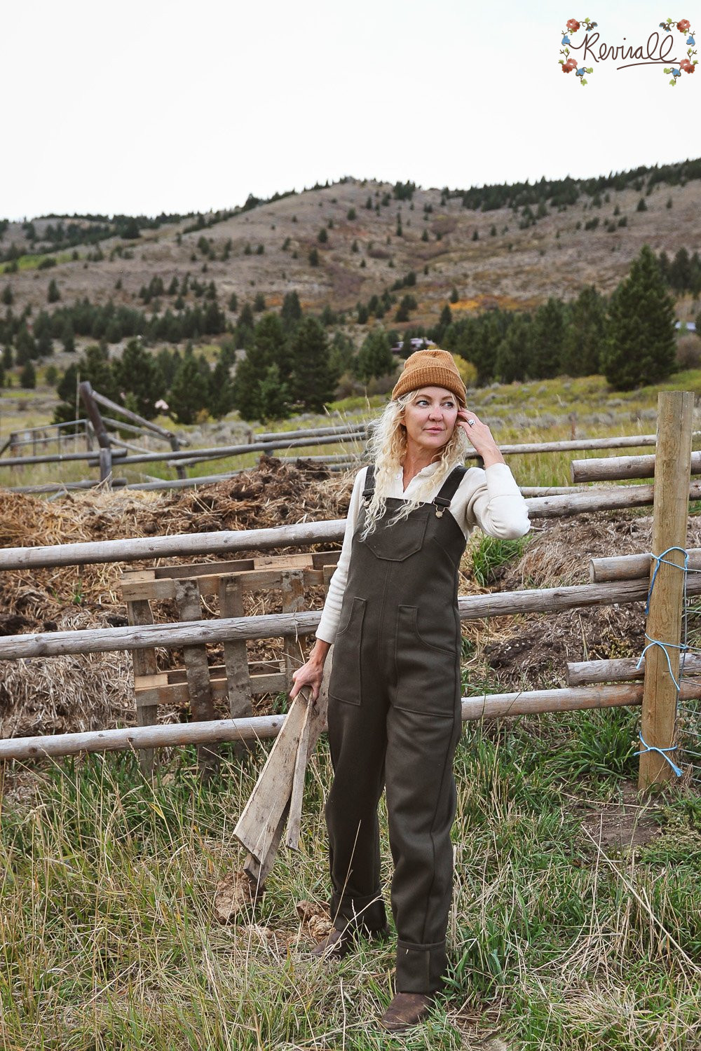 a woman wearing green wool overalls carries a pallet with a wooden fence line and mountains behind her.