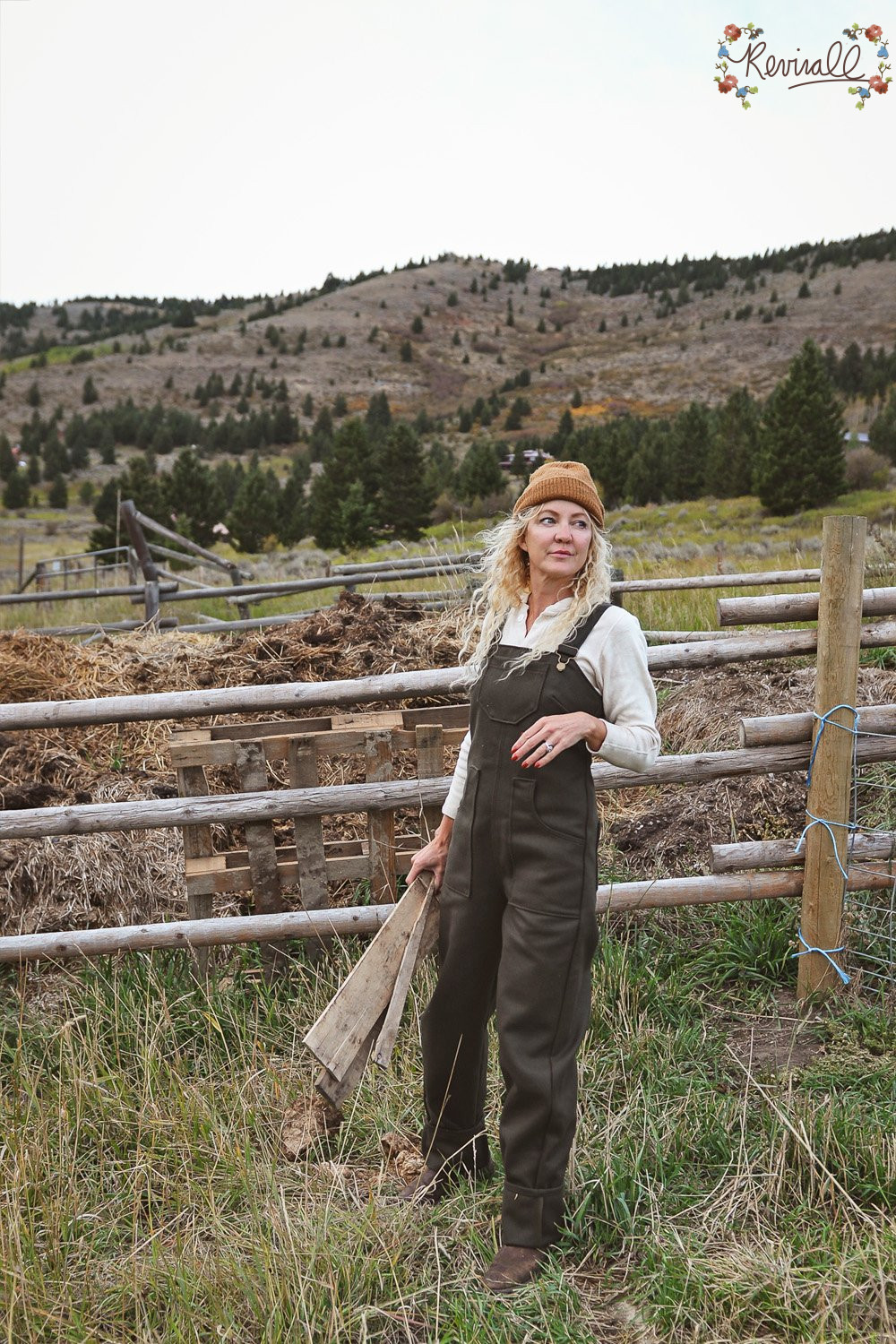 a woman wearing green wool overalls carries a pallet with a wooden fence line and mountains behind her.