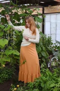 a woman wearing a mustard color cotton skirt with ruffle and thermal shirt stand in a greenhouse next to a large squash plant