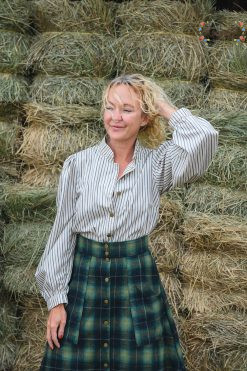 a woman wearing a striped prairie blouse and plaid skirt stands in front of a stack of hay bales